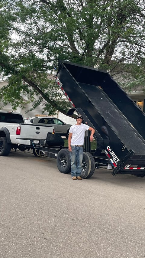 Man standing next to a black dump trailer with raised bed in a driveway