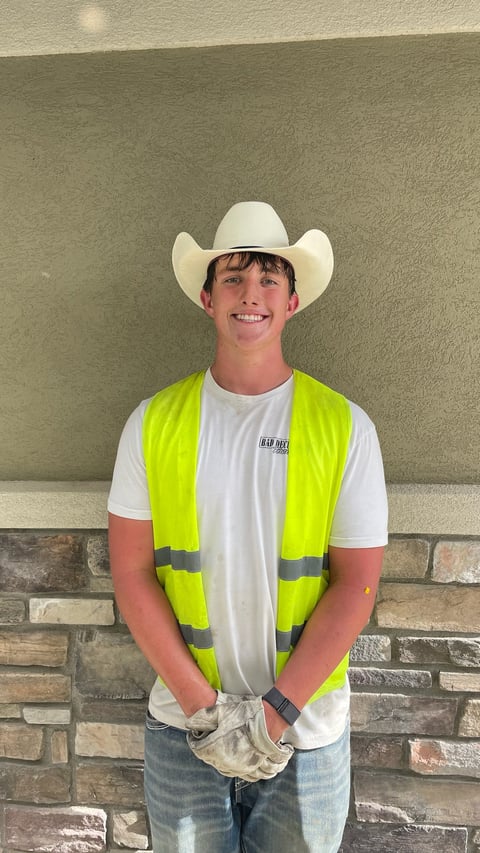 Young man in white cowboy hat, neon yellow safety vest, and white t-shirt smiling in front of a textured wall
