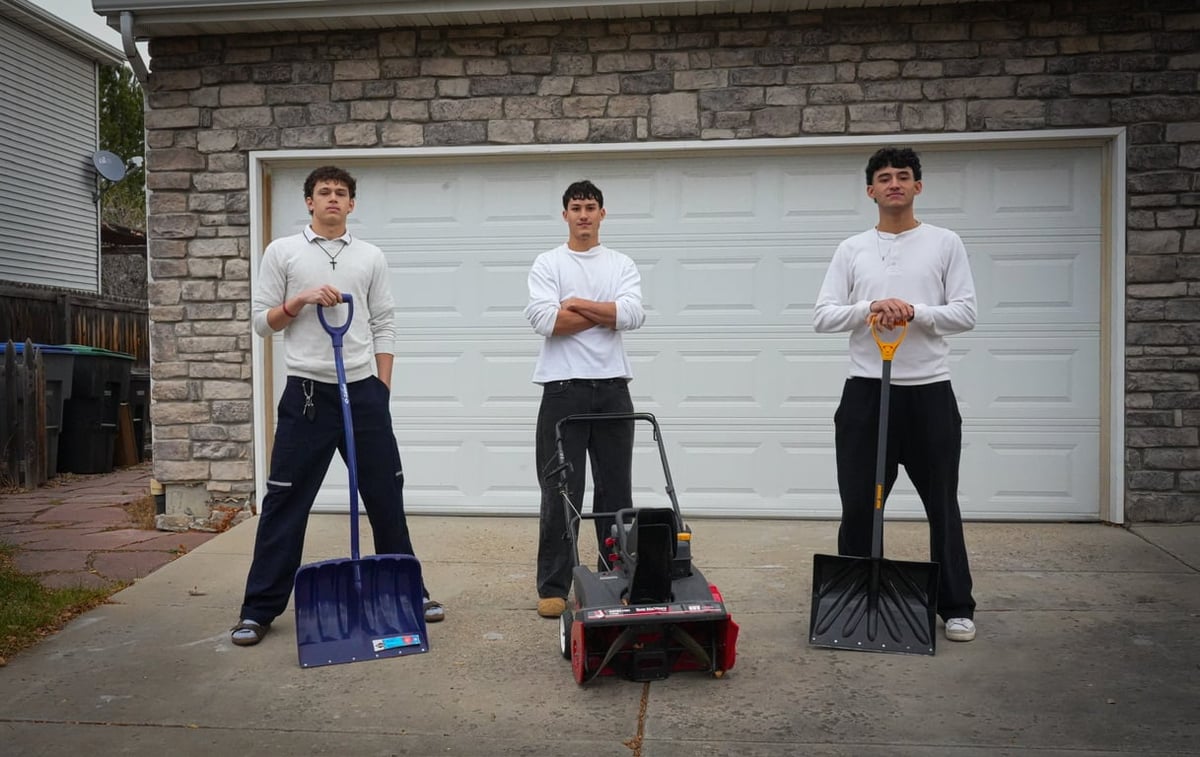 Three men standing in a driveway holding snow shovels in front of a brick house and white garage door
