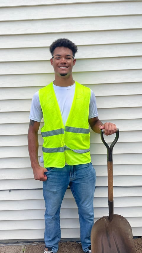 Smiling man wearing neon yellow safety vest holding a shovel against a white siding wall