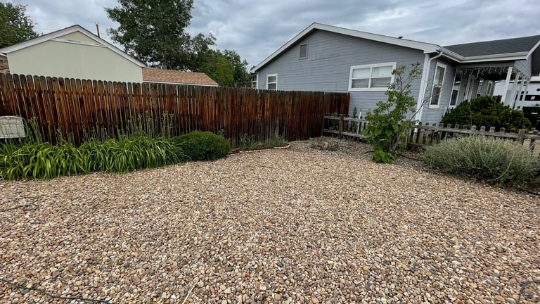 Gravel backyard between two houses with wooden fence, landscaping, and cloudy sky