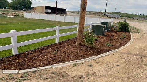 Newly landscaped rural property with white fence, mulched garden bed, and mailbox along dirt driveway