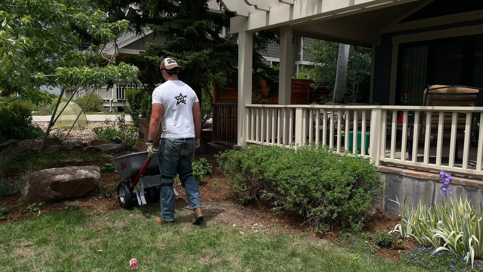 Person in white shirt standing with wheelbarrow in front yard of house with white porch railing