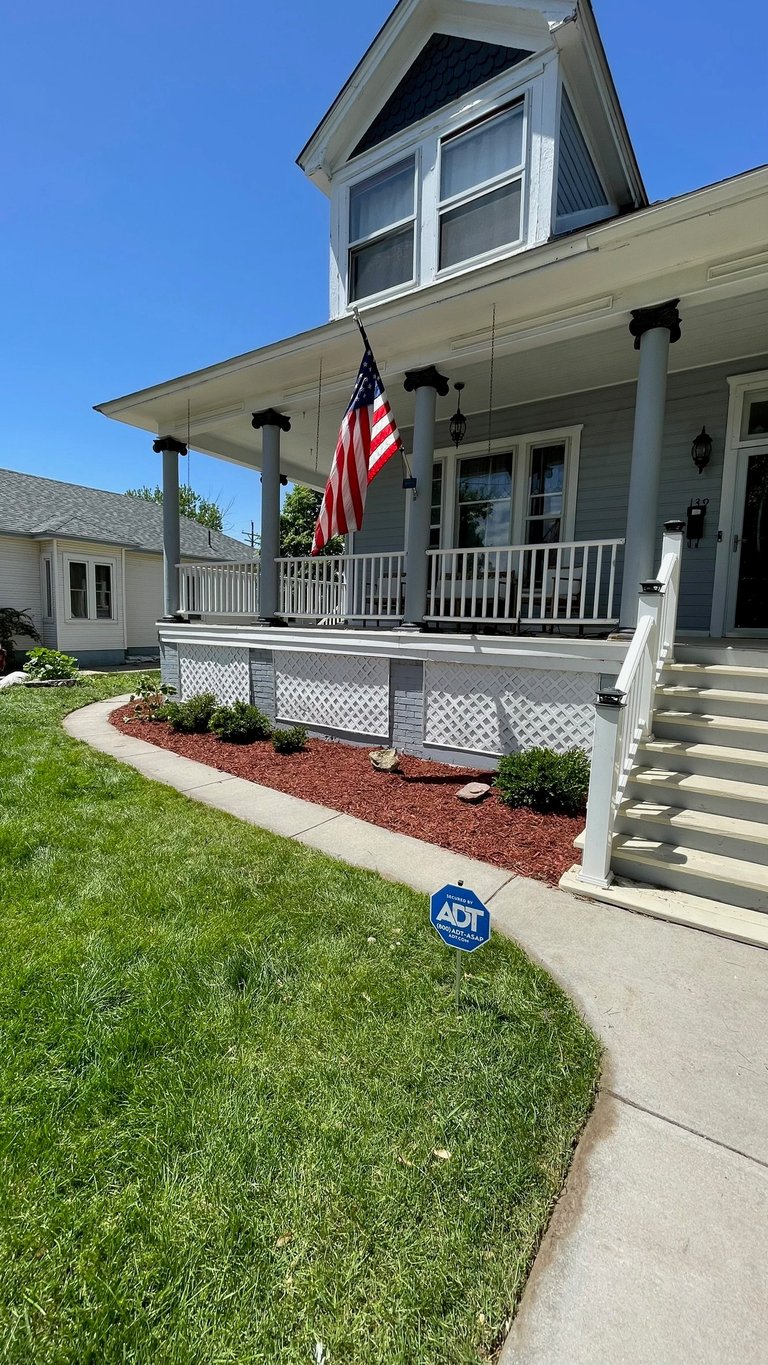 White farmhouse with front porch, black shutters, American flag, lattice skirting, red mulch landscaping, and ADT security sign