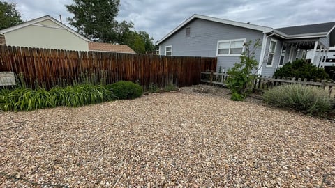 Backyard with gravel ground, wooden fence, and two residential houses under cloudy sky