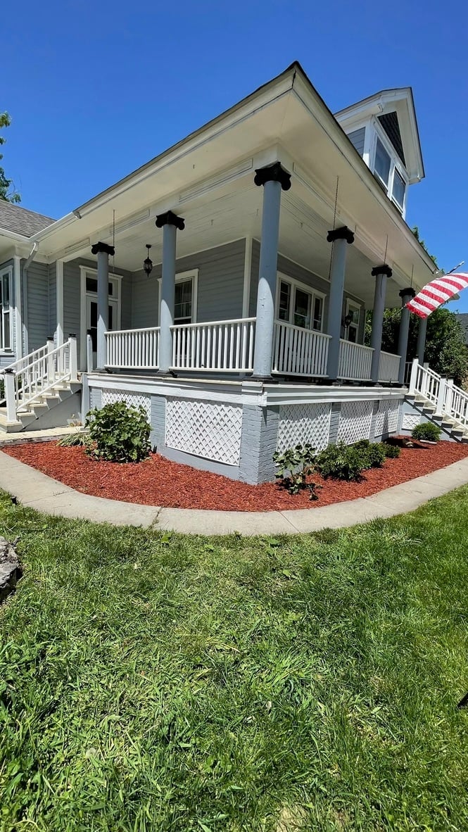 White farmhouse with large front porch, American flag, and lattice skirting under clear blue sky