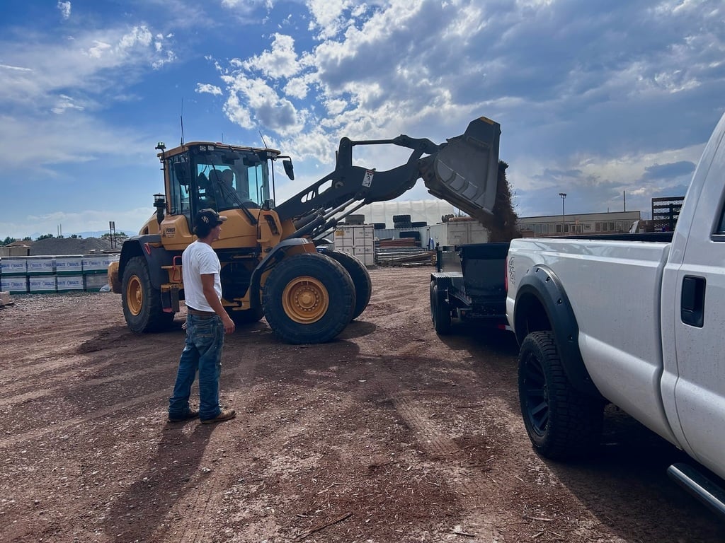Man standing between a yellow wheel loader and white pickup truck on a construction site under blue sky