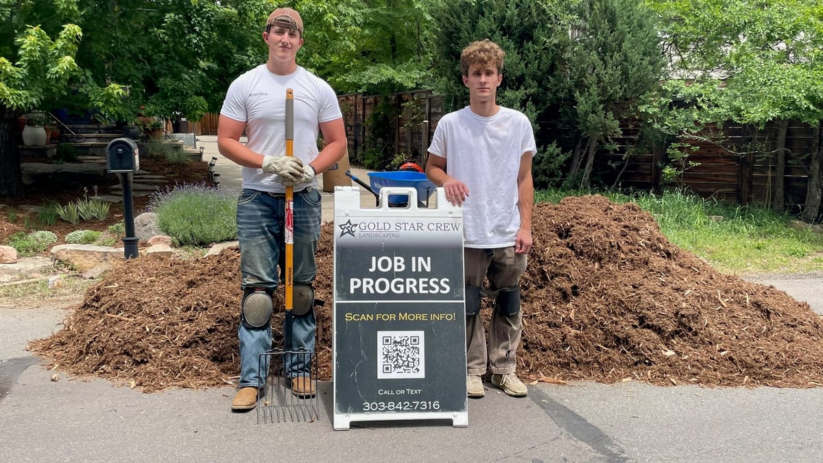 Two people holding a Job In Progress sign while working with mulch piles outdoors, likely documenting a landscaping project.