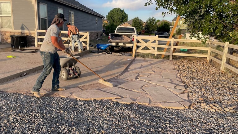 Person operating a concrete saw to cut decorative patterns in a patio or driveway, with wooden fencing and rural property in background