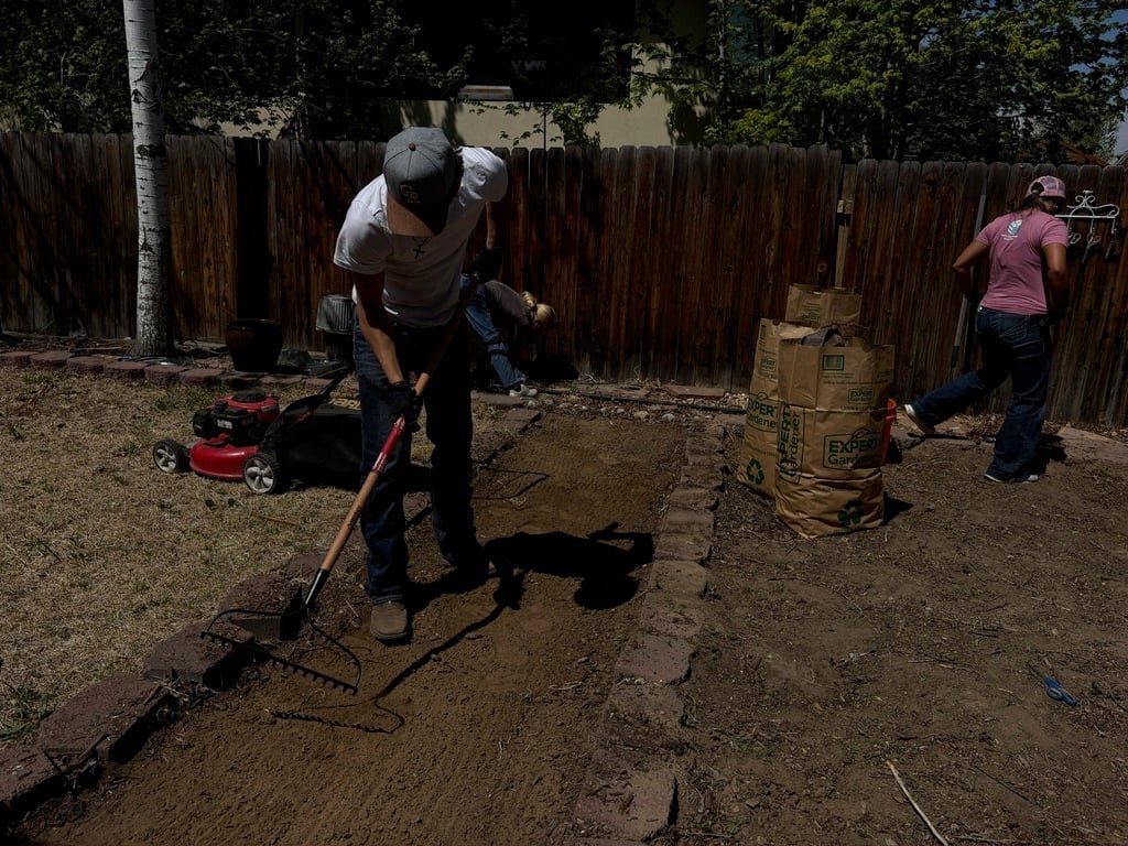 Man in white hard hat using a pickaxe to break up soil in a backyard, with another worker visible in the background