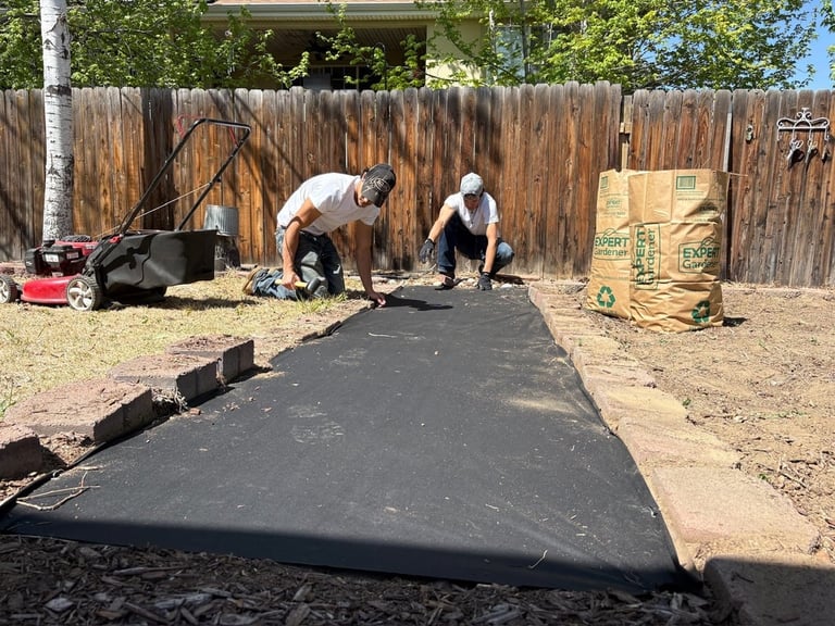 Two workers installing black asphalt or rubber surface in a backyard with wooden fence and landscaping materials visible