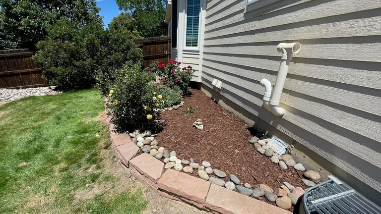 A residential yard with a landscaped flower bed featuring mulch, decorative white stones, and flowering plants along the striped gray siding of a house.