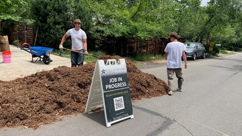 Workers landscaping a residential property with a Job in Progress sign and pile of mulch in the driveway