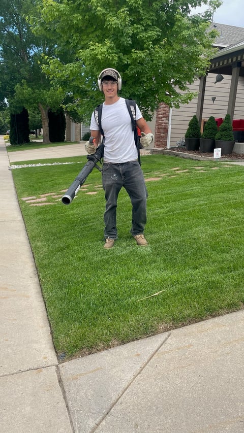 Man wearing headphones and backpack holds a leaf blower while standing on a residential lawn