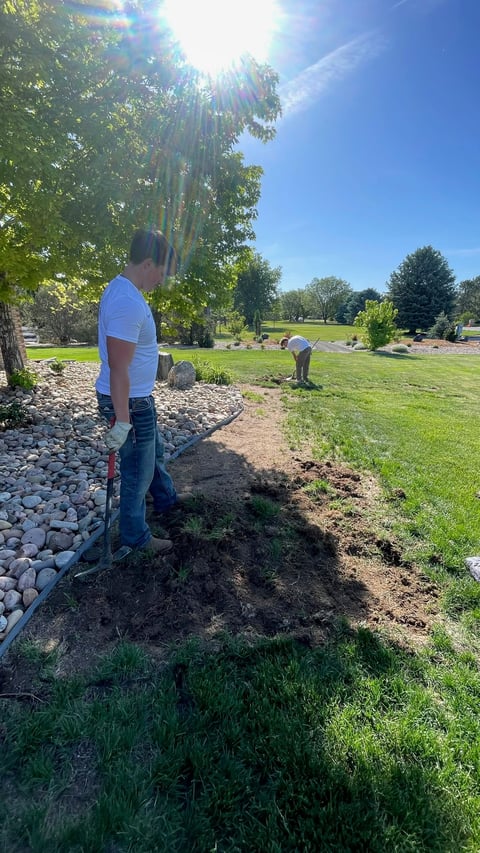 Man digging in dirt at park with shovel on sunny day, trees and green lawn visible in background